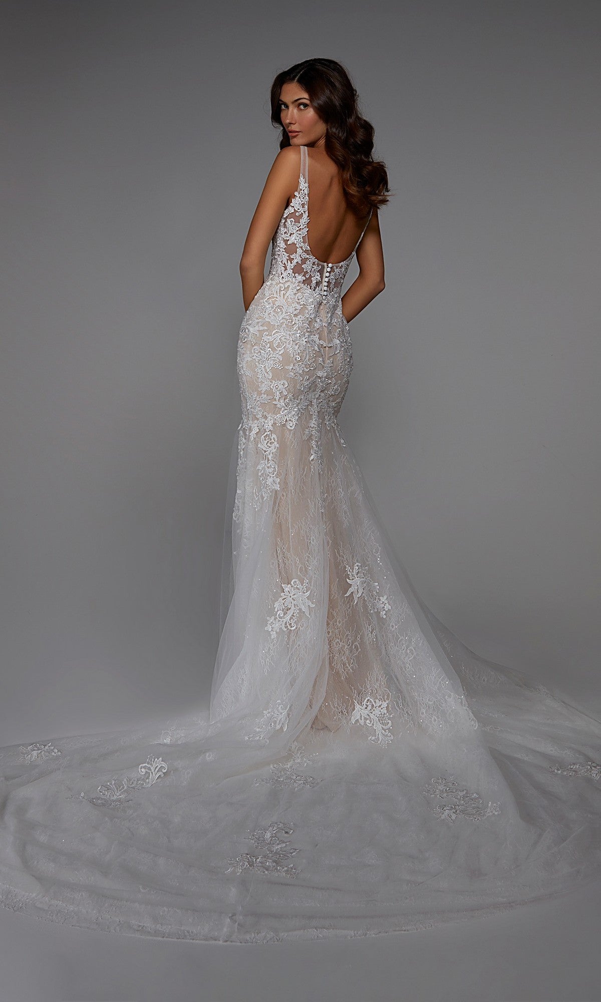 A woman faces away wearing the Alyce 7029, a fitted, backless white lace long formal dress in a trumpet silhouette with an embroidered sheer train, set against a plain gray background.